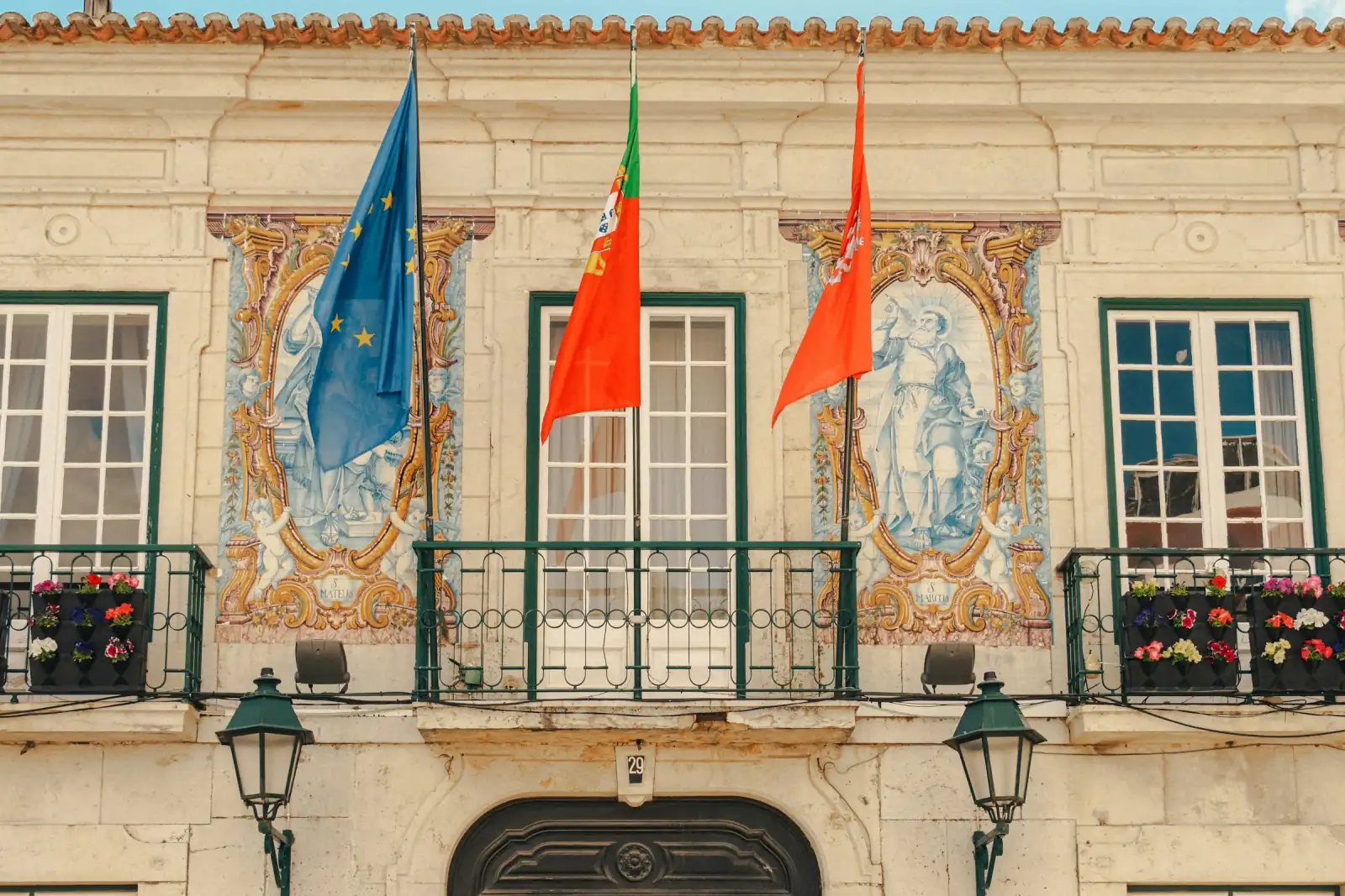 Fachada de um edifício público com as bandeiras de Portugal e da União Europeia, painéis de azulejo e uma varanda de ferro