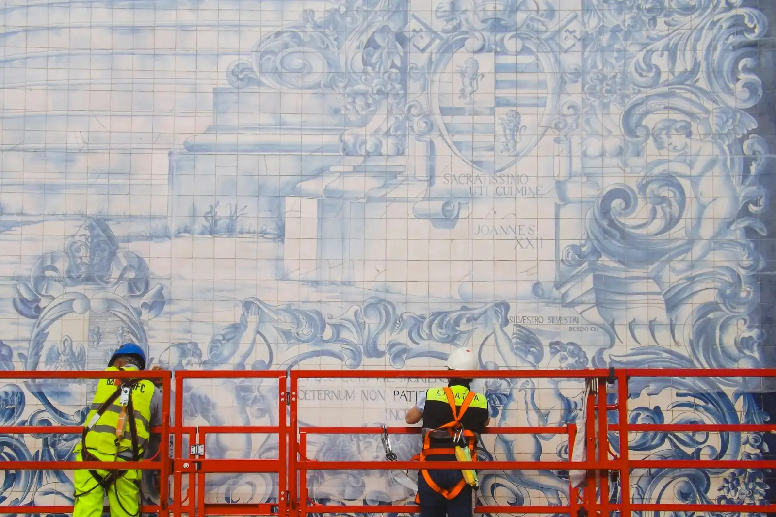 Dois trabalhadores com colete refletor e arnês, numa plataforma elevatória, a restaurar um painel de azulejos azuis e brancos